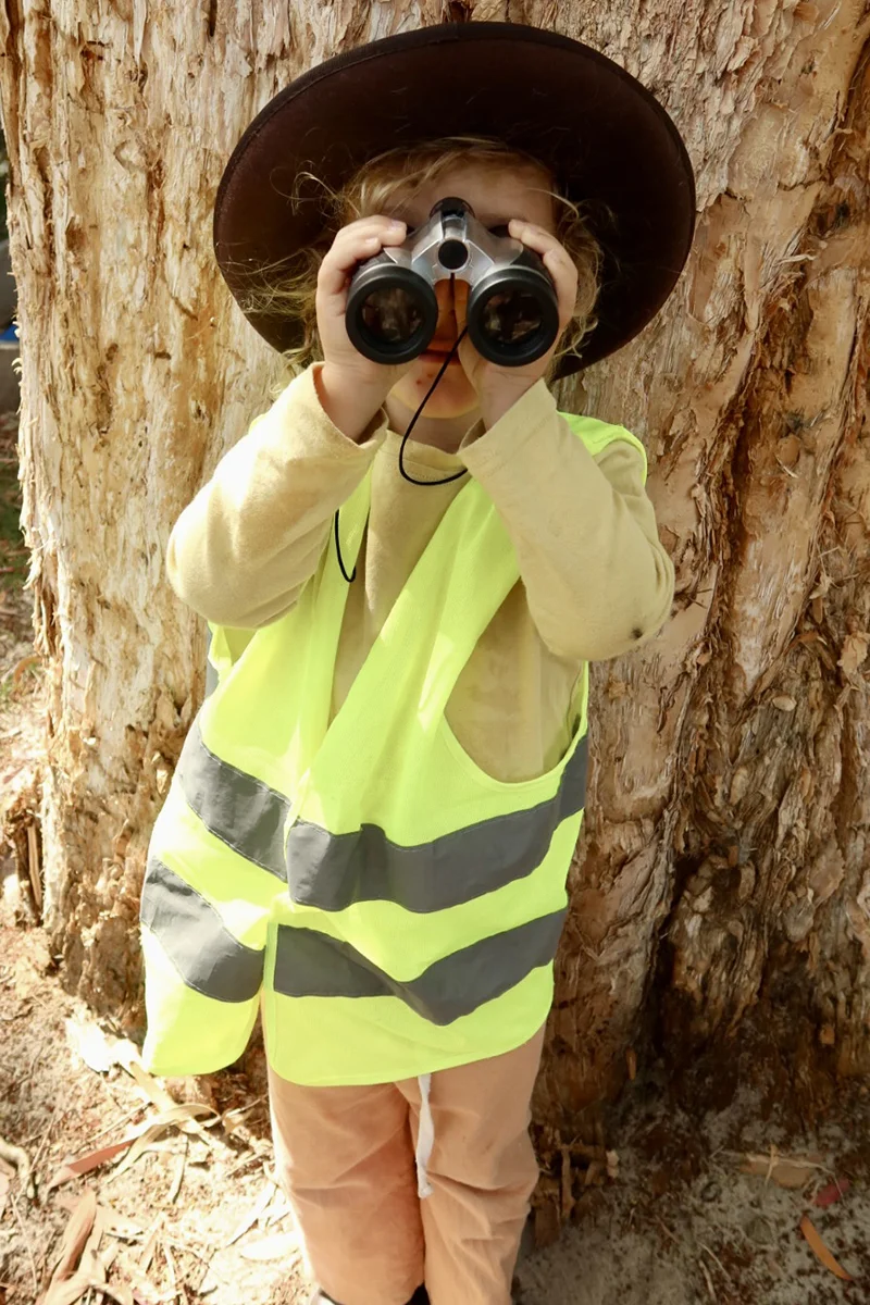 Forest School preschooler in a high-vis vest looking through binoculars during outdoor learning in Tingalpa